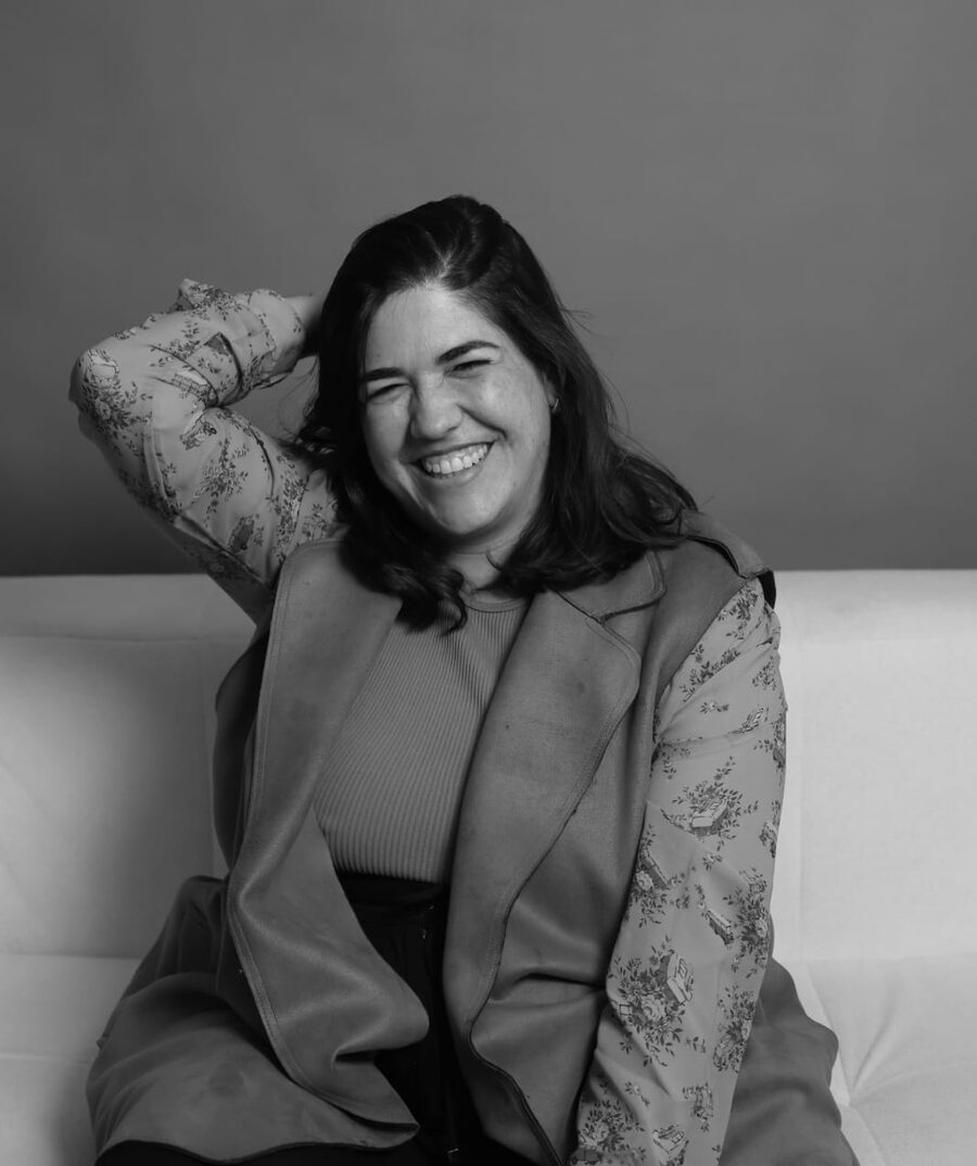 Carol Montenegro sorrindo, sentada, com a mão no cabelo, em foto preto e branco
