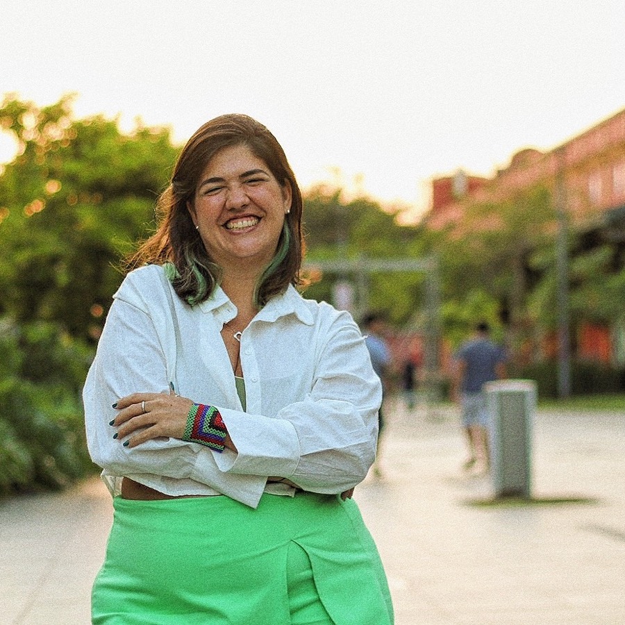 Carol Montenegro sorrindo, de braços cruzados, em um passeio ao ar livre